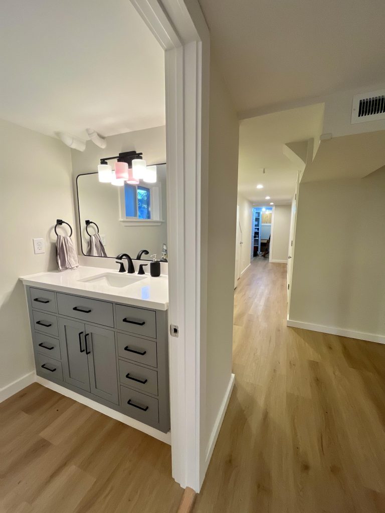 The basement to ADU conversion features a bathroom with a blue vanity before a hallway that extends to the bedrooms and stairs to the main house.