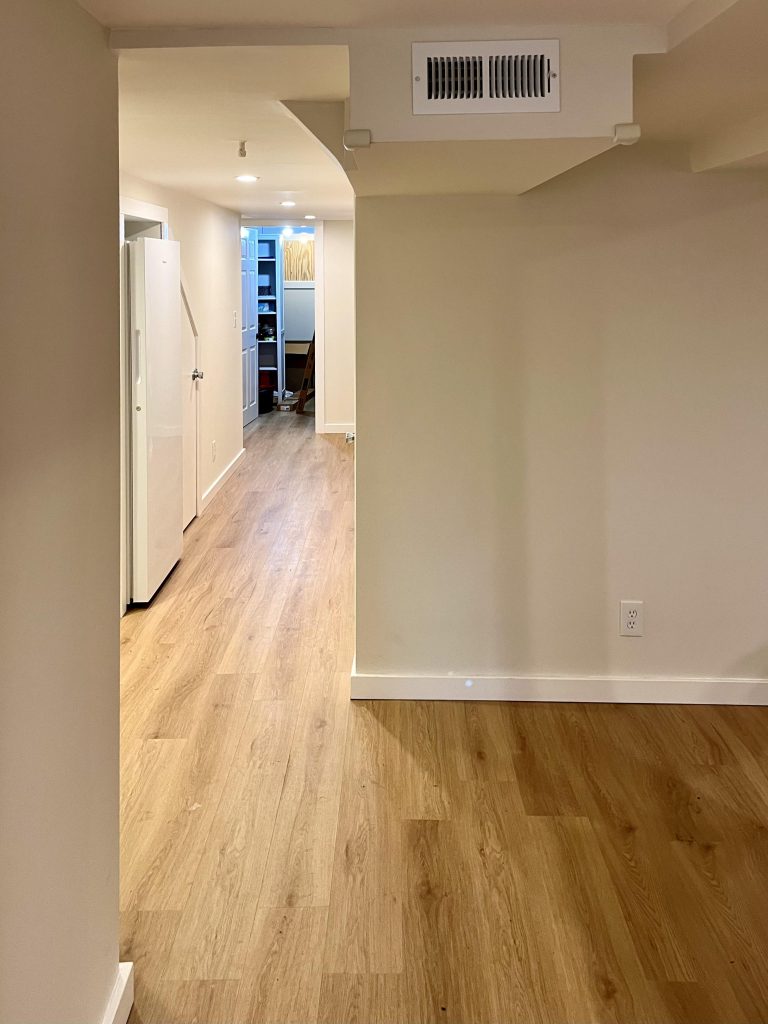 A view from the front to the back of the finished basement shows a refrigerator nook, smooth drywall, and rear storage closet.