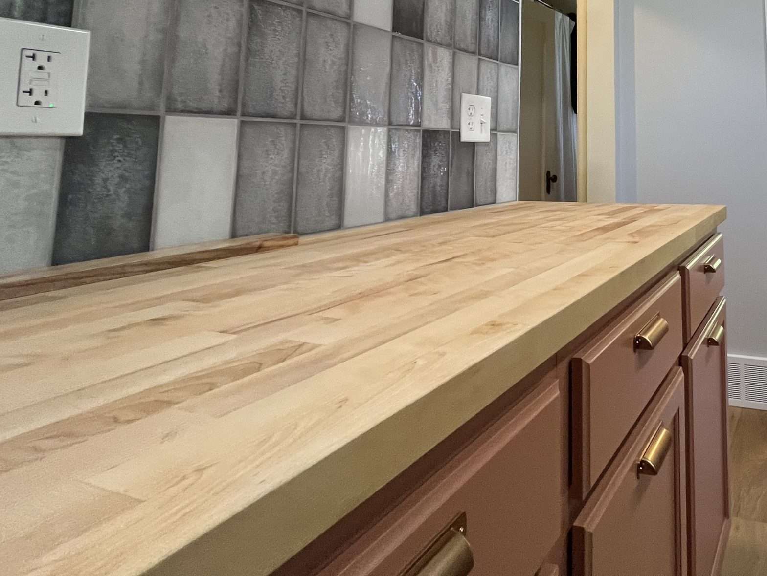 A close-up view of a butcher block countertop with blue-gray tile backsplash and salmon color cabinets.