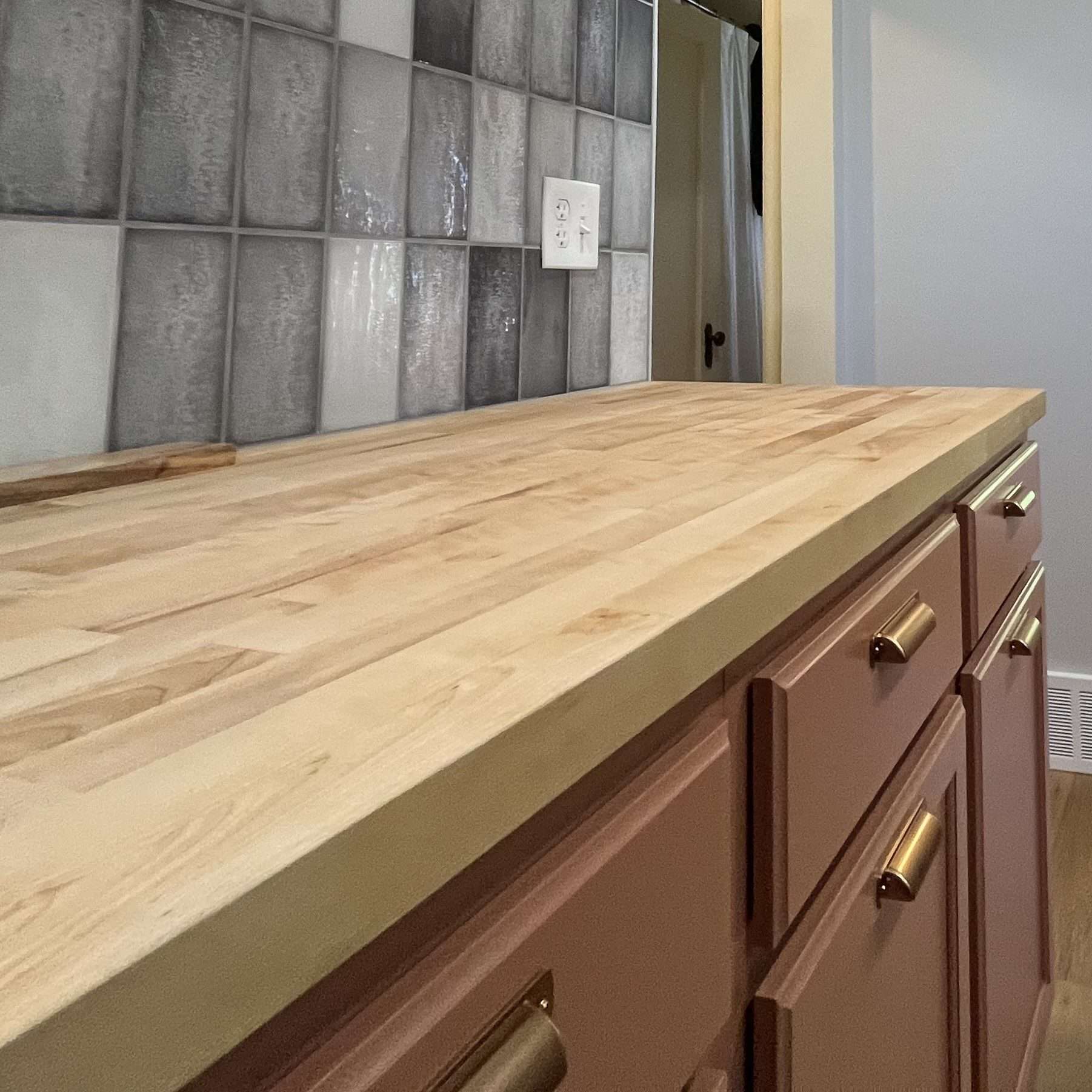 A close-up view of a butcher block countertop with blue-gray tile backsplash and salmon color cabinets.