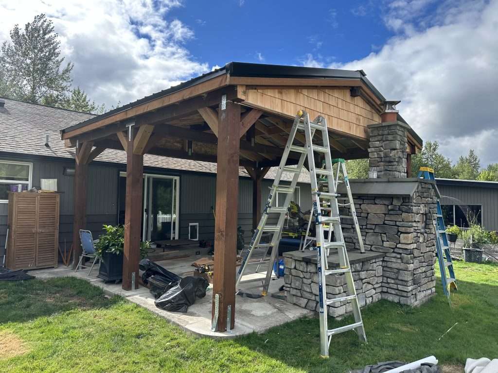 A wide view of a nearly-completed patio cover shows cedar shingles against an existing stone fireplace.