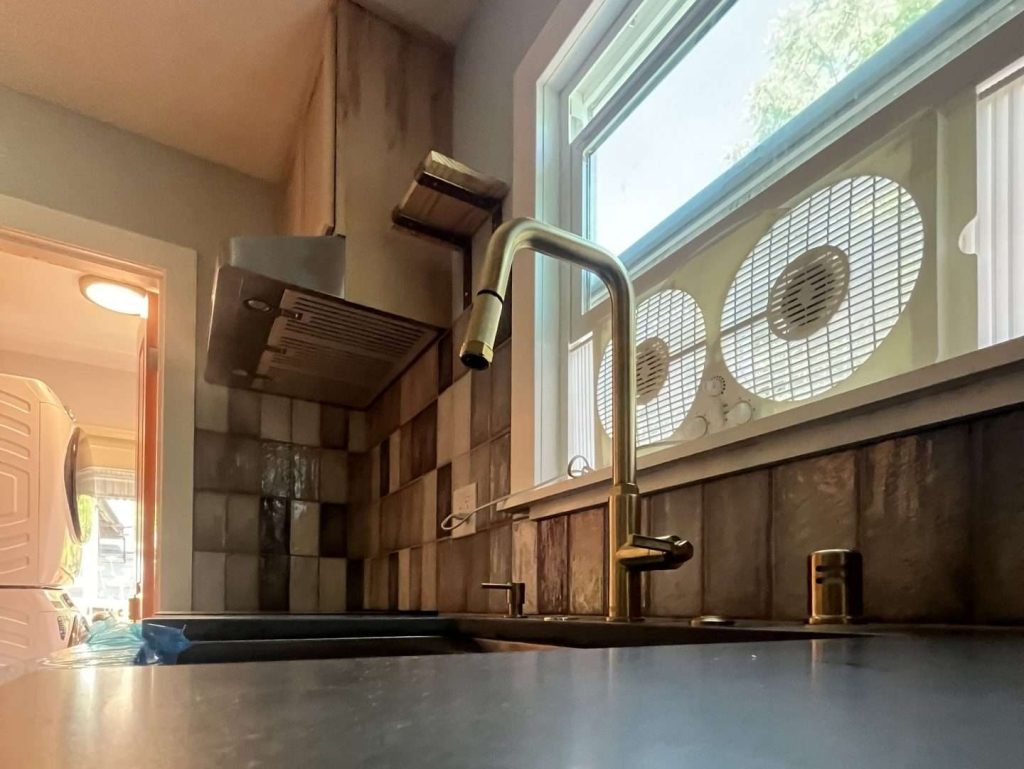 A close up view of a brass kitchen faucet against a dark quartz countertop next to a window with a double fan in it.