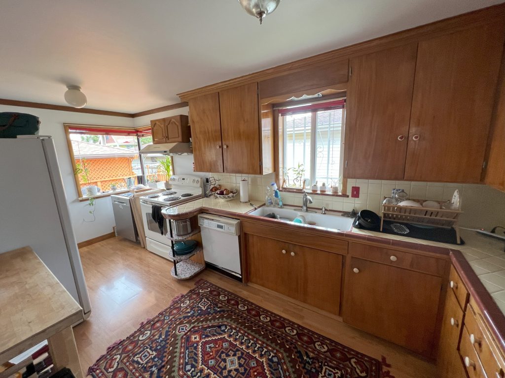 A mid-century-style kitchen has wood cabinets and floors, vinyl counter tops, and dim lighting.