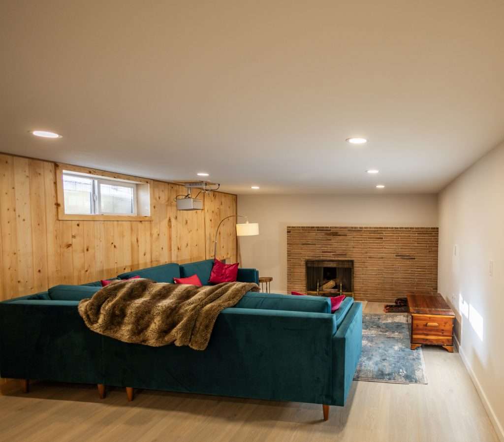 A basement livingroom features a brick fireplace, teal couch, wood-paneled wall, and egress window to allow daylight in.