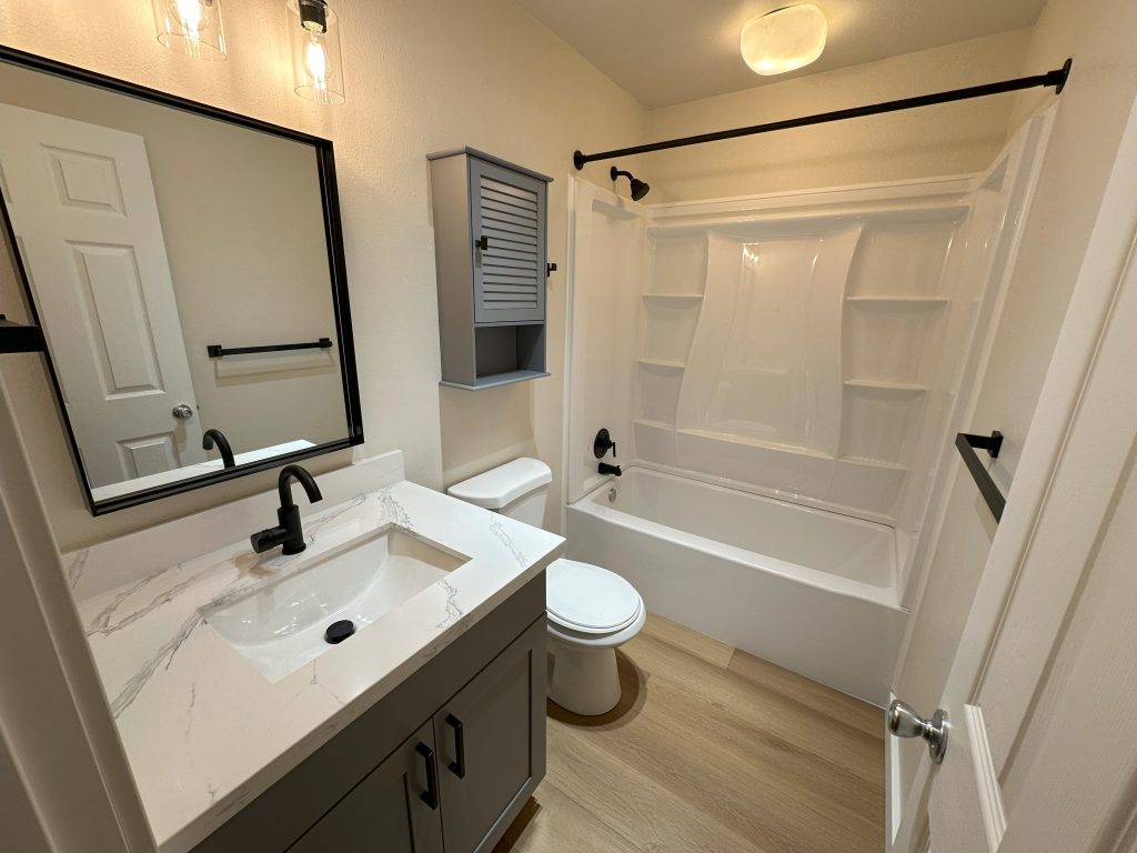 A small bathroom features a vanity with mirror, medicine cabinet, toilet, and bathtub with built-in shelving.