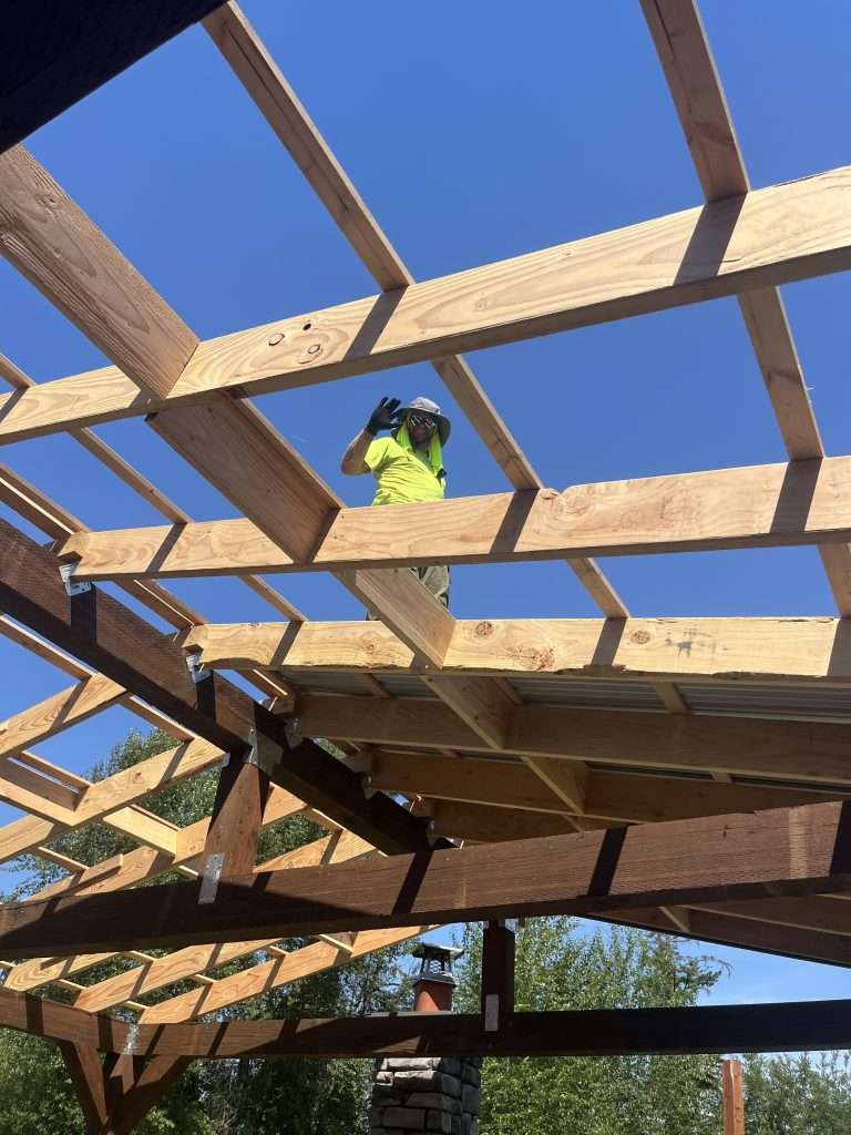 A construction worker waves through the beams as he stands atop the roof framing of a custom patio cover.