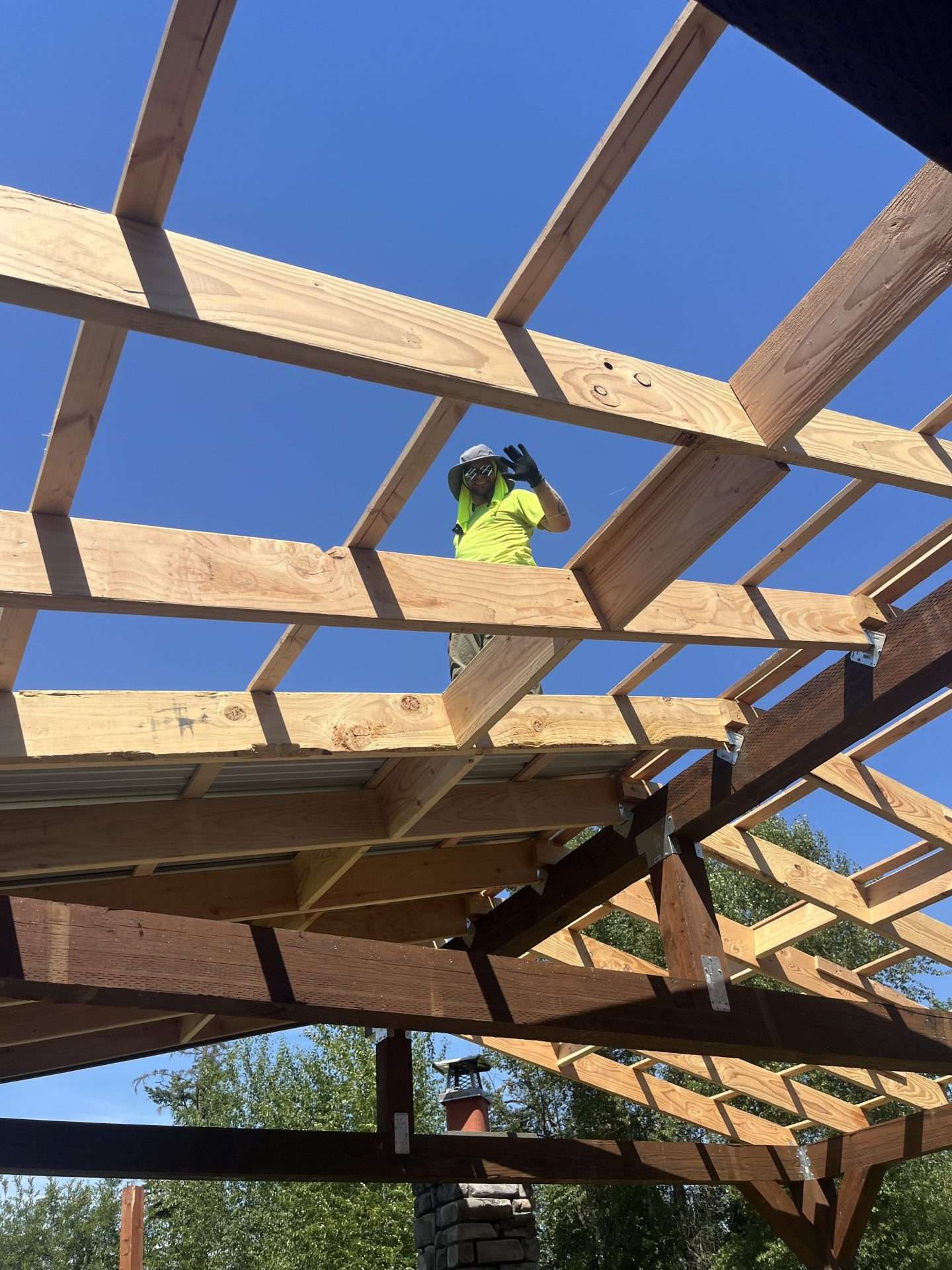 A construction worker waves through the beams as he stands atop the roof framing of a custom patio cover.