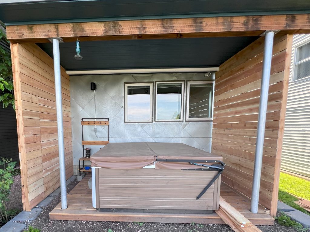 A wooden deck with a roof against the home's metal siding houses a hot tub and towel rack.