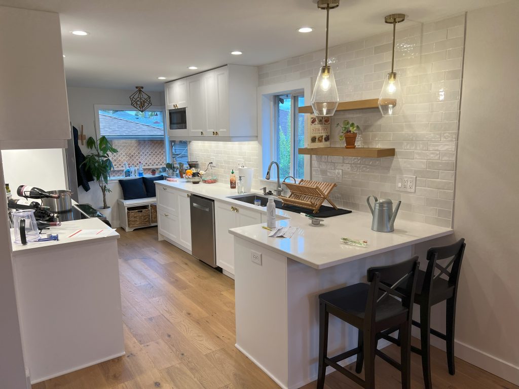 A modern kitchen remodel includes white cabinets, counters, and tiling, as well as bar seating and floating shelves.