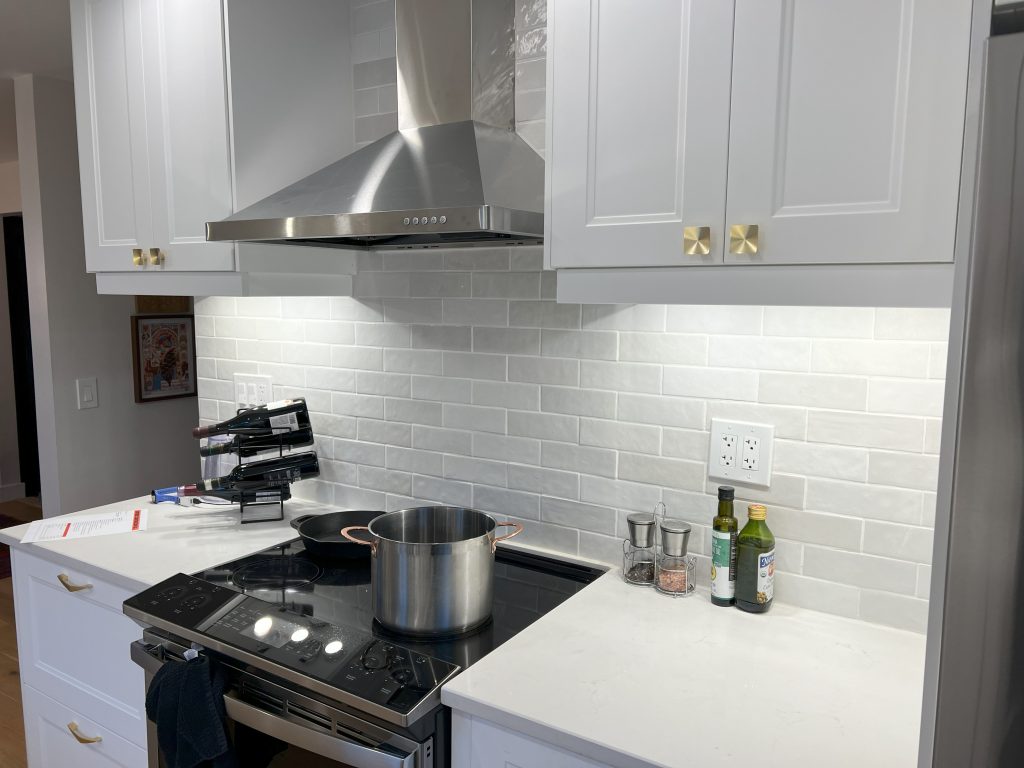 A stovetop with a stainless steel vent, surrounded by white cabinets, counters, and a white tile backsplash.