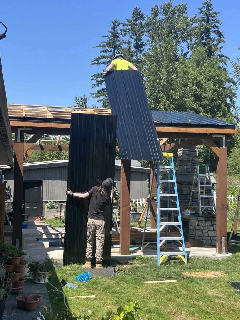 A contractor holds a metal roof panel behind him to carry it onto the roof of the patio cover as a second worker holds another metal panel on the ground beneath.