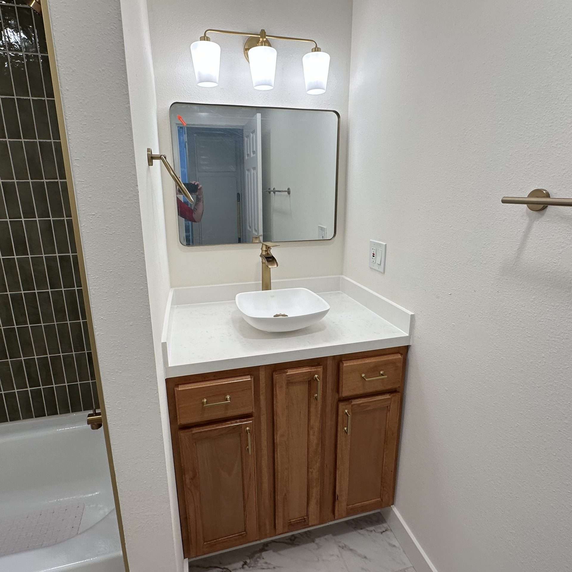 A vanity with three overhead lights illuminatng a vessel sink, next to a bathtub with a green tile surround.