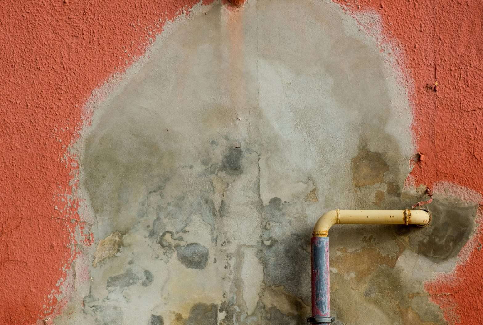 A red wall exposes bare cement and signs of water damage around where pipes come out of it.