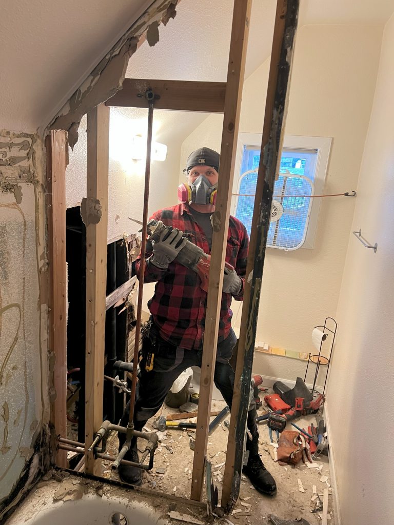 A man stands behind the beams of the former bathtub holding a handsaw and ready to demo the rest of the last of the dividing wall.
